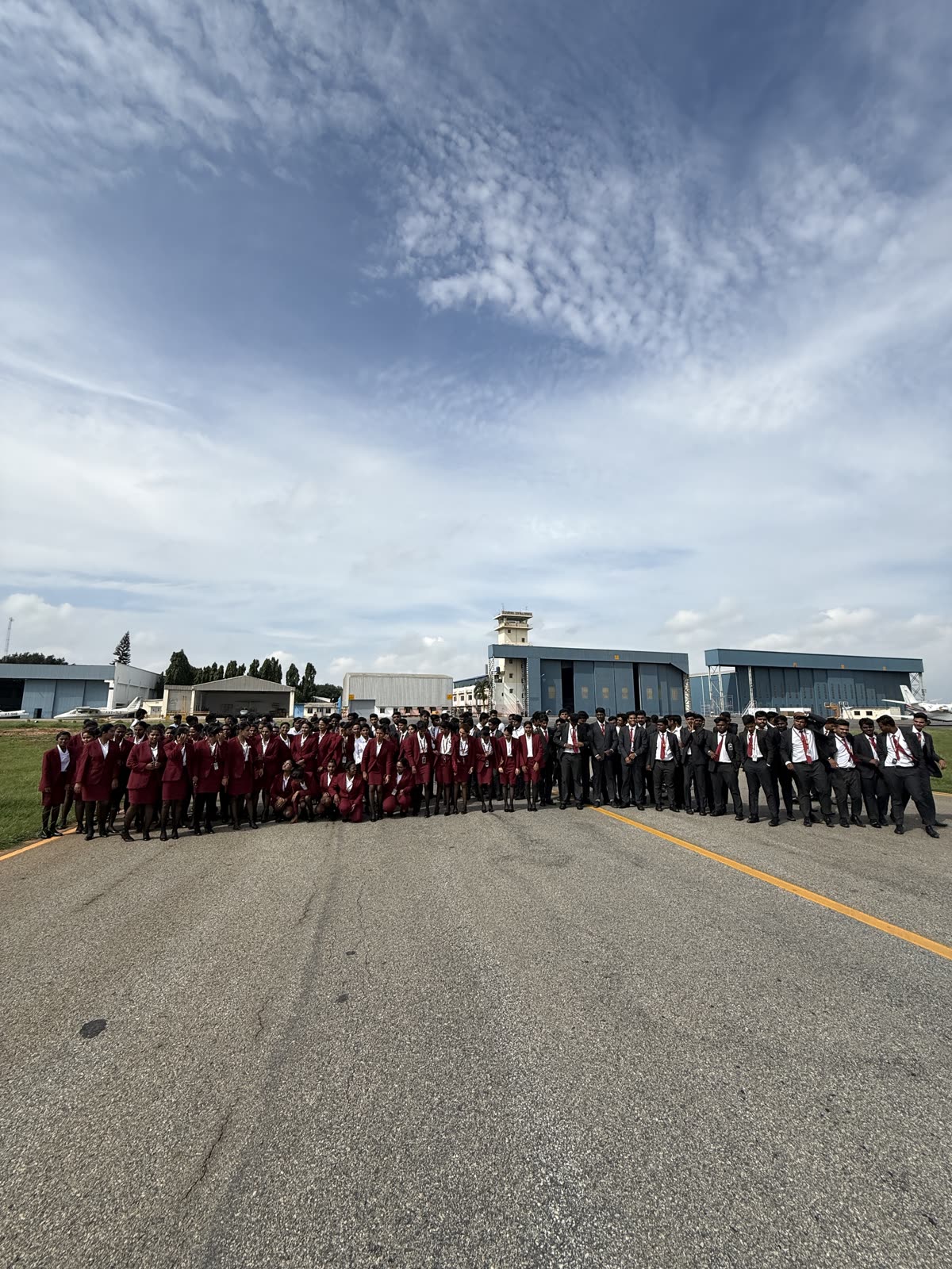 Over a hundred Brandcrew students in uniform standing together on the TAAL airfield taxiway, hangars and tower behind them