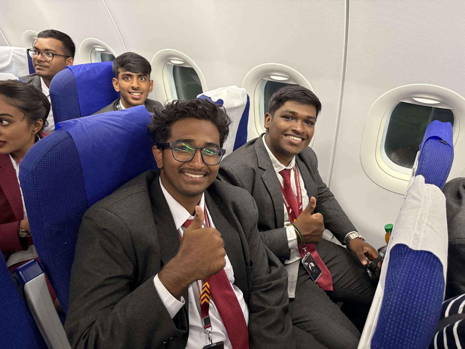Smiling students in suits giving a thumbs up from their seats inside an aircraft cabin